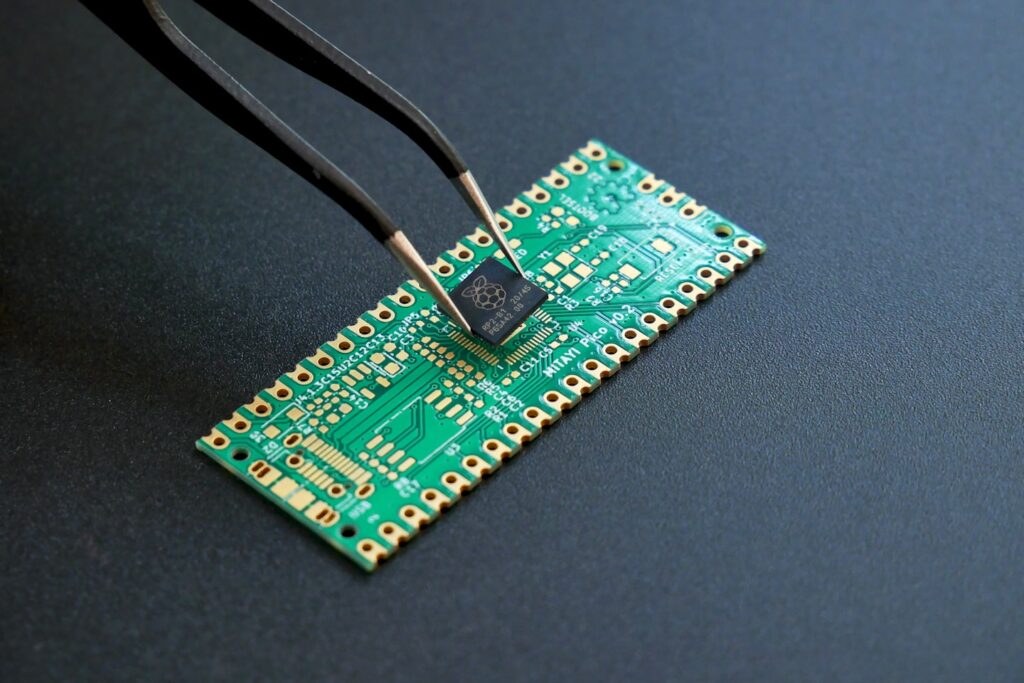 An RP2040 microcontroller held with a tweezer above a PCB. PCB was designed in KiCad and fabricated by PCBWay. Photo shot in Lumix G85.